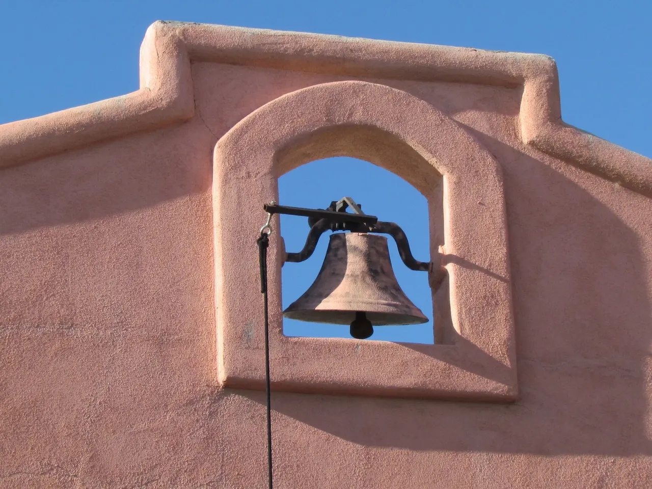 An iron bell set in an arched adobe niche against a deep blue sky
