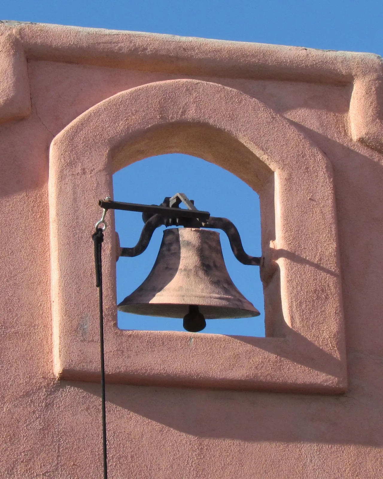 An iron bell set in an arched adobe niche against a deep blue sky